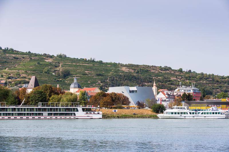 Im Vordergrund ist ein See zu sehen. Die Landesgalerie mit ihrer einzigartigen Form ist zwischen Bäumen hindurch in der Ferne zu sehen. Am Ufer des Sees sind zwei Schiffe zuz sehen. Im Hintergrund an einem Hang sind Weinreben zu erkennen.  Im Vordergrund ist ein See zu sehen. Die Landesgalerie mit ihrer einzigartigen Form ist zwischen Bäumen hindurch in der Ferne zu sehen. Am Ufer des Sees sind zwei Schiffe zuz sehen. Im Hintergrund an einem Hang sind Weinreben zu erkennen.