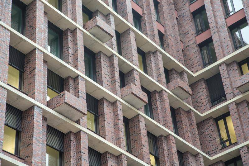 Das Inneneck eines der Gebäude ist zu sehen. Die Gebäudestrucktur ist mit roten und vereinzel schwarzen Backsteinen verkleidet. Dazwischen sind Fenster mit dunkelgrauen Rahmen eingebracht. Schwarze Jalousien sind an den Fenstern angebracht. Das Inneneck eines der Gebäude ist zu sehen. Die Gebäudestrucktur ist mit roten und vereinzel schwarzen Backsteinen verkleidet. Dazwischen sind Fenster mit dunkelgrauen Rahmen eingebracht. Schwarze Jalousien sind an den Fenstern angebracht.