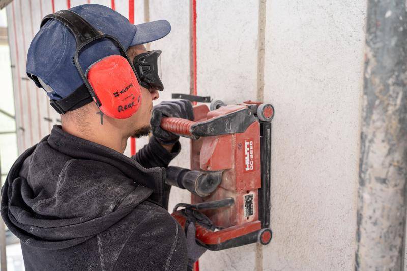 Ein Arbeiter mit rotem Gehörschutz und Schutzbrille fräst mit einer roten Maschine der Firma Hilti die Fugen für die Leitungen in die alte, verputzte Fassade. Im hintergrund sind bereits rote Leitungen in der Wand zu sehen. Ein Arbeiter mit rotem Gehörschutz und Schutzbrille fräst mit einer roten Maschine der Firma Hilti die Fugen für die Leitungen in die alte, verputzte Fassade. Im hintergrund sind bereits rote Leitungen in der Wand zu sehen.