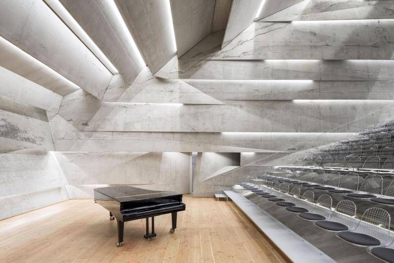 Innenansicht des Konzertsaals. Auf der Bühne mit Holzbelag steht ein schwarzer Flügel. Recht steigt die Tribüne mit den Sitzlätzen an. Die Innenwände sind aus Sichtbetonelementen hergestellt, die durch indirekte Beleuchtung mit LED-Lichtern unterbrochen sind. Innenansicht des Konzertsaals. Auf der Bühne mit Holzbelag steht ein schwarzer Flügel. Recht steigt die Tribüne mit den Sitzlätzen an. Die Innenwände sind aus Sichtbetonelementen hergestellt, die durch indirekte Beleuchtung mit LED-Lichtern unterbrochen sind.