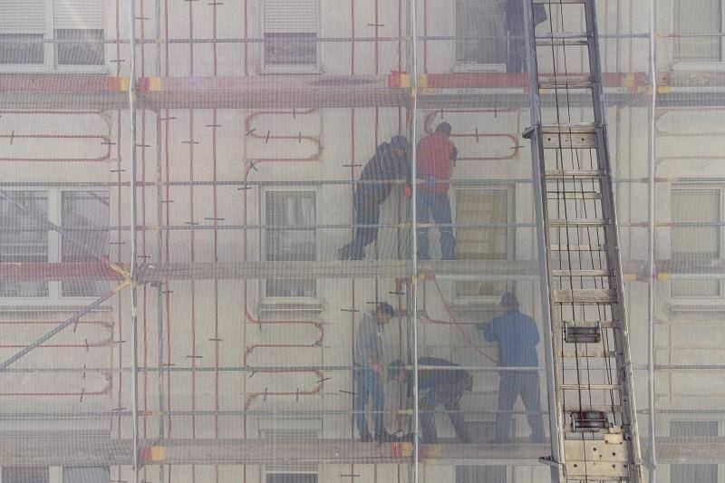 Hinter den Schutznetzen des Gerüsts sind Arbeiter zu sehen, die zwischen den Fenstern die roten Leitungen verlegen und besfestigen. Hinter den Schutznetzen des Gerüsts sind Arbeiter zu sehen, die zwischen den Fenstern die roten Leitungen verlegen und besfestigen.