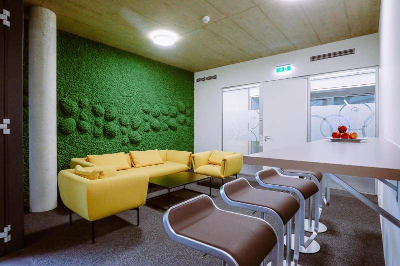 Aufenthaltsraum mit gelben Sitzmöbeln, sowie einer grünen, bemoosten Wand dahinter. Aufenthaltsraum mit gelben Sitzmöbeln, sowie einer grünen, bemoosten Wand dahinter.