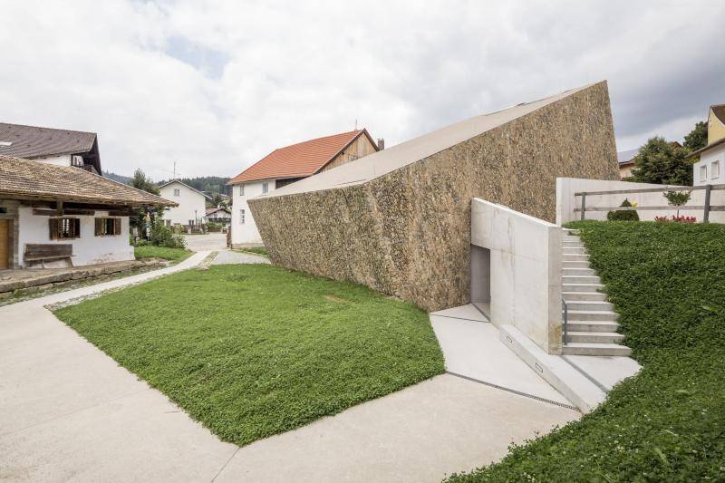 Die Hinterseite des Gebäudes ist zu sehen. Daneben ein eingeschossiges altes Wohnhaus mit Holz-Läden. Die schräge Dachfläche des Gebäudes ist zu sehen. Rechts am Gebäude befindet sich eine Treppe aus Sichtbeton, die hinauf zum höhergelegenen Dorfplatz führt. Die Hinterseite des Gebäudes ist zu sehen. Daneben ein eingeschossiges altes Wohnhaus mit Holz-Läden. Die schräge Dachfläche des Gebäudes ist zu sehen. Rechts am Gebäude befindet sich eine Treppe aus Sichtbeton, die hinauf zum höhergelegenen Dorfplatz führt.