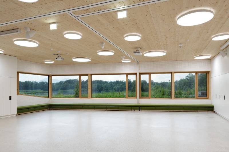 Aufnahme aus dem Inneren des Gebäude. Die aneinander gereihten Fenster lassen den See und den Wald draußen wie ein Panoramabild wirken. Der Raum ist Hell, groß und leer. Unter den Fenstern ist eine lange, grüne Sitzbank. Aufnahme aus dem Inneren des Gebäude. Die aneinander gereihten Fenster lassen den See und den Wald draußen wie ein Panoramabild wirken. Der Raum ist Hell, groß und leer. Unter den Fenstern ist eine lange, grüne Sitzbank.