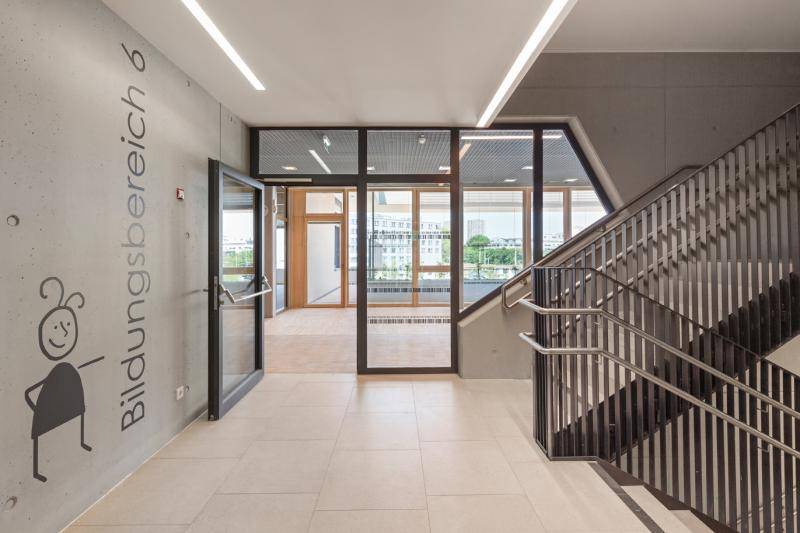 Ansicht aus dem Treppenhaus. Die Wände sind aus Sichtbeton. Das Treppengeläder aus Metall in anthrazit. Eine große Glastüre öffnet den Bereich hinter dem Treppenhaus. Ansicht aus dem Treppenhaus. Die Wände sind aus Sichtbeton. Das Treppengeläder aus Metall in anthrazit. Eine große Glastüre öffnet den Bereich hinter dem Treppenhaus.