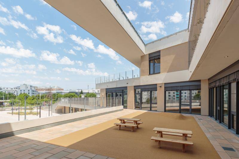 Freibereich im ersten Obergeschoss. Zwei Picknicktische aus Holz stehen dort. Die Oberfläche ist in dunklem Gelb. Im Hintergrund öffnet sich der Blick über das Geländer auf andere Gebäude der Stadt und auf den begrünten Vorplatz des Gebäudes. Freibereich im ersten Obergeschoss. Zwei Picknicktische aus Holz stehen dort. Die Oberfläche ist in dunklem Gelb. Im Hintergrund öffnet sich der Blick über das Geländer auf andere Gebäude der Stadt und auf den begrünten Vorplatz des Gebäudes.
