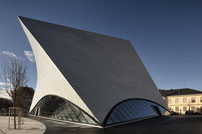 Die Schräggeneigte Fassade des Gebäudes ist in voller größe zu sehen. Unten ist ein Halbkreis aus Glas angeordnet. Die linke Seite des Gebäudes, die nach oben spitz zuläuft, ist von der Sonne angetrahlt. Die Schräggeneigte Fassade des Gebäudes ist in voller größe zu sehen. Unten ist ein Halbkreis aus Glas angeordnet. Die linke Seite des Gebäudes, die nach oben spitz zuläuft, ist von der Sonne angetrahlt.