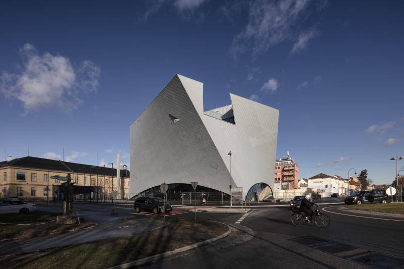Das Gebäude ist hinter einer Straße zu sehen. Auf der Straße fahren Autos und ein Fahrrad. Oben am Gebäude ist ein Ausschnitt mit einem Glasgeländer zu sehen. Dort befindet sich eine Dachterrasse. Das Gebäude ist hinter einer Straße zu sehen. Auf der Straße fahren Autos und ein Fahrrad. Oben am Gebäude ist ein Ausschnitt mit einem Glasgeländer zu sehen. Dort befindet sich eine Dachterrasse.