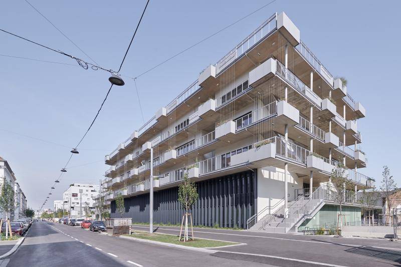 Frontansicht des Gebäudes. Die ersten zwei Stockwerke haben eine durchlaufende Fassade in anthrazit. Neben dem Gebäude führt eine Treppe im Freien zum Eingang im 1.OG. Die oberen Geschosse, sowie die Dachterrasse haben hellgraue Geländer und weiße aussenwände, sowie Sichtbeton Untersichten an den Balkonen. Frontansicht des Gebäudes. Die ersten zwei Stockwerke haben eine durchlaufende Fassade in anthrazit. Neben dem Gebäude führt eine Treppe im Freien zum Eingang im 1.OG. Die oberen Geschosse, sowie die Dachterrasse haben hellgraue Geländer und weiße aussenwände, sowie Sichtbeton Untersichten an den Balkonen.