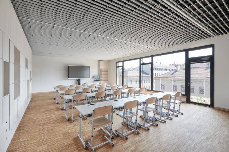 Innenaufnahme in einem Unterrichtsraum. Der Boden mit hellem Stäbchenparkett, weiße Wände, große Fensterfläche mit grauen Fensterrahmen. Tischreihen mit Stühlen in Richtung Smart-Board ausgerichtet. Decke mit weißen senkrecht stehenden Lamellen verkleidet. Innenaufnahme in einem Unterrichtsraum. Der Boden mit hellem Stäbchenparkett, weiße Wände, große Fensterfläche mit grauen Fensterrahmen. Tischreihen mit Stühlen in Richtung Smart-Board ausgerichtet. Decke mit weißen senkrecht stehenden Lamellen verkleidet.
