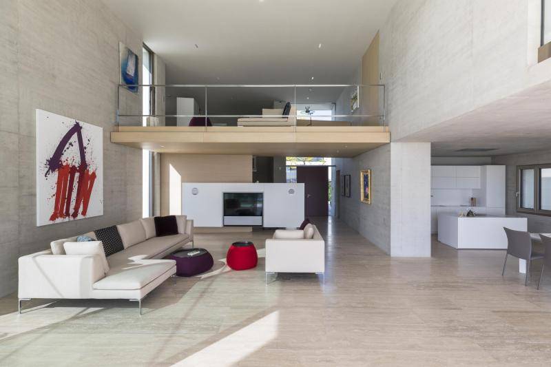 Aufnahme aus dem Inneren der Villa. Zu sehen ist der Wohnbereich im Zentrum mit zwei Sofas in beige. Rechts daneben ist teilweise die offene Küche zu sehen. Das Wohnzimmer ist nach oben offen und auf der Galerie ist ein weiteres kleines Sofa zu sehen. An den Sichtbetonwänden hängen diverse Gemälde. Aufnahme aus dem Inneren der Villa. Zu sehen ist der Wohnbereich im Zentrum mit zwei Sofas in beige. Rechts daneben ist teilweise die offene Küche zu sehen. Das Wohnzimmer ist nach oben offen und auf der Galerie ist ein weiteres kleines Sofa zu sehen. An den Sichtbetonwänden hängen diverse Gemälde.