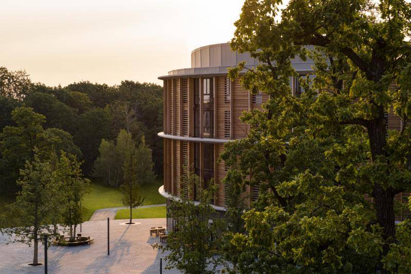 Das Gebäude ist durch einen Baum verdeckt, doch die Holzfassade ist durch das Grün zu sehen. Links ist die Terrasse vor dem runden Gebäude zu sehen Das Gebäude ist durch einen Baum verdeckt, doch die Holzfassade ist durch das Grün zu sehen. Links ist die Terrasse vor dem runden Gebäude zu sehen