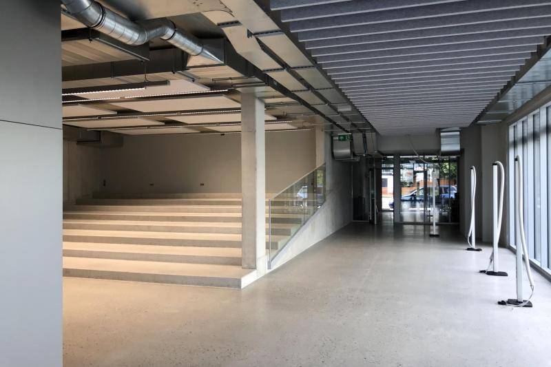 Der Eingangsbereich des Gebäudes. Im linken Bildteil ist eine Tribüne mit Stufen aus Beton angeordnet. An der Decke sind sichtbare Installationen, rechts ist eine Glasfassade nach außen zu sehen. Der Eingangsbereich des Gebäudes. Im linken Bildteil ist eine Tribüne mit Stufen aus Beton angeordnet. An der Decke sind sichtbare Installationen, rechts ist eine Glasfassade nach außen zu sehen.
