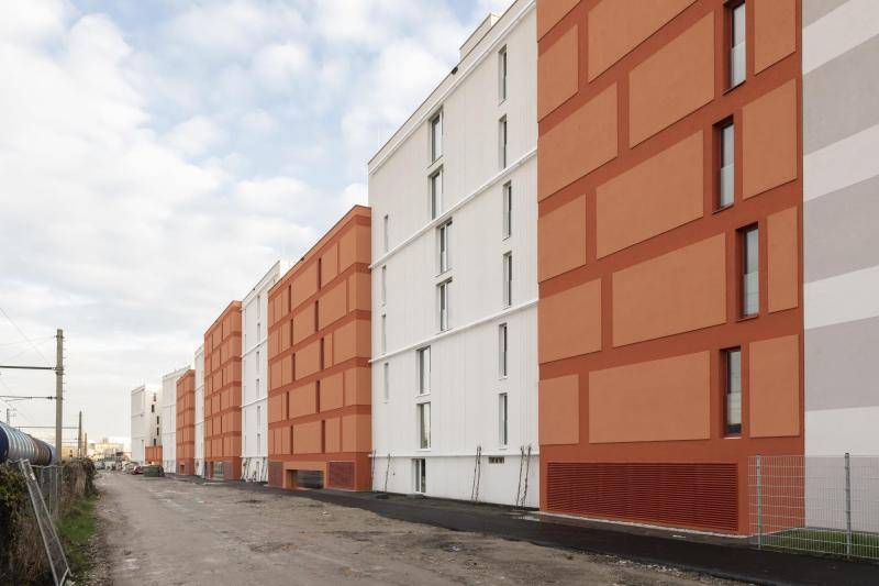 Der Hauptgebäudeteil ist in voller Länge zu sehen, davor verläuft eine Bau/SchotterStraße. Die Gebäudefassade ist im Wechsel mit je 10m Länge, orange und weiß. Vereinzelt sind rechteckige Fenster angeordnet. Der Hauptgebäudeteil ist in voller Länge zu sehen, davor verläuft eine Bau/SchotterStraße. Die Gebäudefassade ist im Wechsel mit je 10m Länge, orange und weiß. Vereinzelt sind rechteckige Fenster angeordnet.