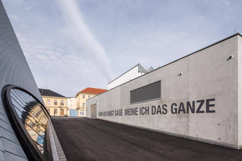 Hier ist auf einer Sichtbetonfassade der Spruch "Wenn ich Kunst sage meine ich das Ganze" geschrieben. Links neben einem kleinen Hof steigt dann die Fassade der Galeria in die Höhe. Im Hintergrund sind andere Gebäude zu sehen. Hier ist auf einer Sichtbetonfassade der Spruch "Wenn ich Kunst sage meine ich das Ganze" geschrieben. Links neben einem kleinen Hof steigt dann die Fassade der Galeria in die Höhe. Im Hintergrund sind andere Gebäude zu sehen.
