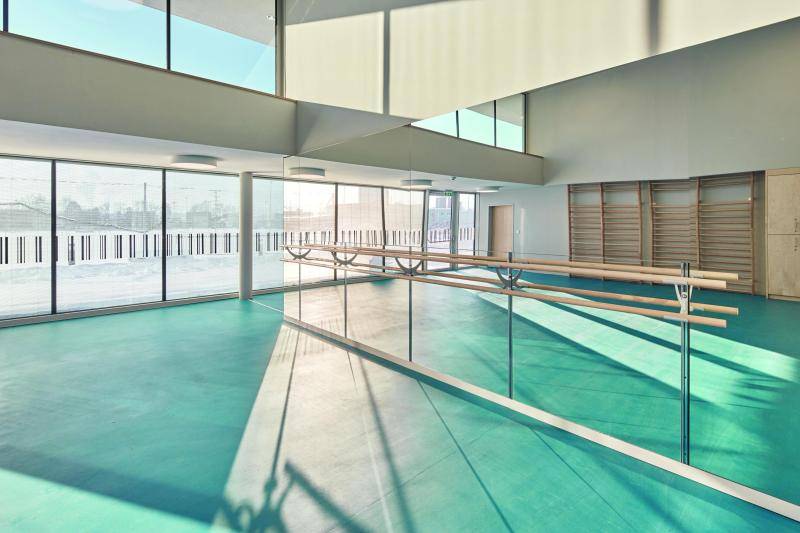 Hier sieht man einen Turnraum der Halle. An einer Spielgelwand sind Holzstangen agebracht. Im Spiegel sieht man Kletterwände aus Holz. Links ist eine Fensterfront nach draußen, durch die man eine verschneite Landschaft sieht. Der Hallenboden leutet türkis durch das Tageslicht. Hier sieht man einen Turnraum der Halle. An einer Spielgelwand sind Holzstangen agebracht. Im Spiegel sieht man Kletterwände aus Holz. Links ist eine Fensterfront nach draußen, durch die man eine verschneite Landschaft sieht. Der Hallenboden leutet türkis durch das Tageslicht.