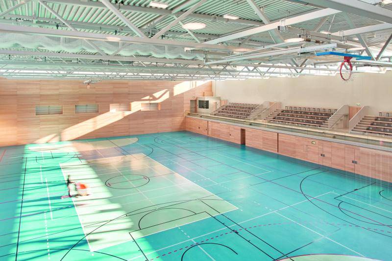 Der Blick von oben in die große Sporthalle. Die Wände aus Holz, ebenso die kleine Tribüne auf der rechten Seite. Die Dachkonstruktion sind weiße Fachwerkträger. Auf dem Hallenboden sind viele Linien für Spielfelder verschiedenster Sportarten eingezeichnet. Der Blick von oben in die große Sporthalle. Die Wände aus Holz, ebenso die kleine Tribüne auf der rechten Seite. Die Dachkonstruktion sind weiße Fachwerkträger. Auf dem Hallenboden sind viele Linien für Spielfelder verschiedenster Sportarten eingezeichnet.