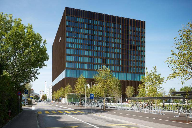 An der Fassade schimmern die großen Glasflächen türkis. Unterbrohen werden die Scheiben durch braune, metall Stäbe, die die Fassade gestalten. Das vierte Geschoss ist vollverglast, sodass die darüberliegenden Geschosse wie schwebend wirken. An der Fassade schimmern die großen Glasflächen türkis. Unterbrohen werden die Scheiben durch braune, metall Stäbe, die die Fassade gestalten. Das vierte Geschoss ist vollverglast, sodass die darüberliegenden Geschosse wie schwebend wirken.