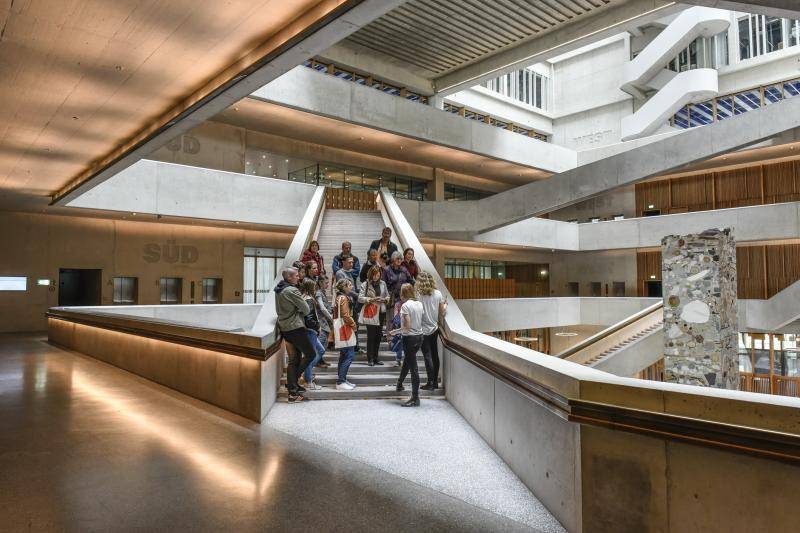 Eine Gruppe von Besucher*innen steht auf einer der Treppen im großen Foyerraum. Die Geländer sind aus Sichtbeton mit Holzhandläufen. Eine Gruppe von Besucher*innen steht auf einer der Treppen im großen Foyerraum. Die Geländer sind aus Sichtbeton mit Holzhandläufen.