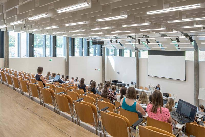 Zu sehen ist ein Hörsaal mit Sitzplätzen als Tribühne angeordnet. Die Sitze sind aus Holz. Einige Menschen sitzen im Raum. Vorn steht eine Lehrperson vor einer Leinwand. Duch Fenster kommt viel natürliches Licht in den Raum. An der Decke hängen weiße, rechteckige Akkustik-Elemente und Lichtröhren. Zu sehen ist ein Hörsaal mit Sitzplätzen als Tribühne angeordnet. Die Sitze sind aus Holz. Einige Menschen sitzen im Raum. Vorn steht eine Lehrperson vor einer Leinwand. Duch Fenster kommt viel natürliches Licht in den Raum. An der Decke hängen weiße, rechteckige Akkustik-Elemente und Lichtröhren.