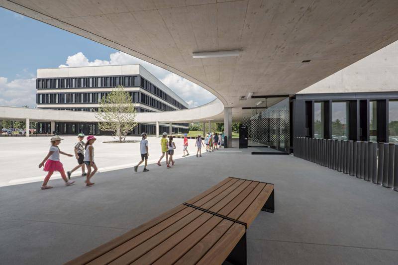 Zu sehen der Hof zwischen den Gebäuden mit der runden Überdachung in Form eines Halbkreises. Die Überdachung ist auch Sichtbeton. Einige Kinder laufen über den Hof. Im Vordergrund steht eine Bank aus Holz, im Hintergrund ist eines der Schulgebäude zu sehen. Zu sehen der Hof zwischen den Gebäuden mit der runden Überdachung in Form eines Halbkreises. Die Überdachung ist auch Sichtbeton. Einige Kinder laufen über den Hof. Im Vordergrund steht eine Bank aus Holz, im Hintergrund ist eines der Schulgebäude zu sehen.