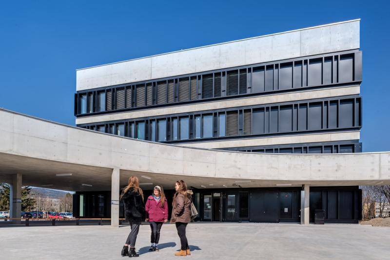 Zu sehen die Überdachung im Hof. Dahinter ist eines der Gebäude der Schule zu sehen. Dessen Fassade besteht aus Festern in schwarzen Rahmen und Beton. Auf dem Hof stehen drei junge Frauen. Zu sehen die Überdachung im Hof. Dahinter ist eines der Gebäude der Schule zu sehen. Dessen Fassade besteht aus Festern in schwarzen Rahmen und Beton. Auf dem Hof stehen drei junge Frauen.