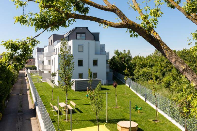Blick aus der Distanz auf die Stirnseite des Hauses. Im Gartenteil sind Tisch und Bänke, sowie einige Spielgeräte. Junge Bäume wurden angepflanzt. Blick aus der Distanz auf die Stirnseite des Hauses. Im Gartenteil sind Tisch und Bänke, sowie einige Spielgeräte. Junge Bäume wurden angepflanzt.