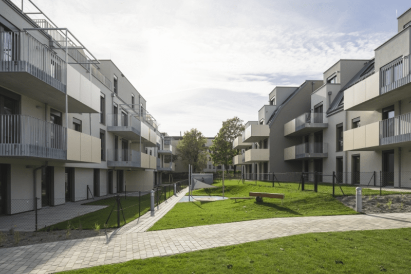 Links und rechts ist je eines der Gebäude zu sehen. Zahlreiche Balkone ragen in den Zwischenraum. Dort führt ein schmaler Weg zwischen den Gebäuden hindurch, sowie eine kleine Sandgrube ist mit Bänken dort plaziert. Links und rechts ist je eines der Gebäude zu sehen. Zahlreiche Balkone ragen in den Zwischenraum. Dort führt ein schmaler Weg zwischen den Gebäuden hindurch, sowie eine kleine Sandgrube ist mit Bänken dort plaziert.