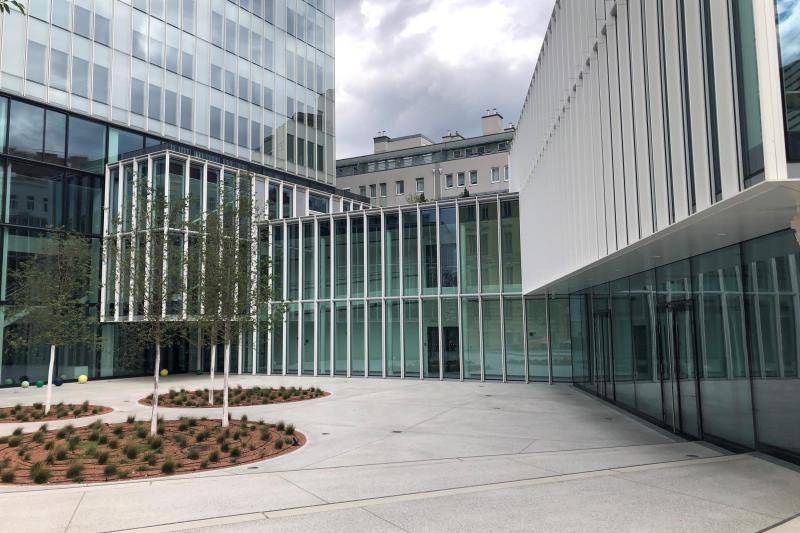Der Vorplatz des Gebäudes mit seinen Hellen Betonplatten und den Beeten. Rechts im Bild der Gebäudeteil mit zwei Geschossen. Links beginnt der Hochhausteil des Komplexes. Der Vorplatz des Gebäudes mit seinen Hellen Betonplatten und den Beeten. Rechts im Bild der Gebäudeteil mit zwei Geschossen. Links beginnt der Hochhausteil des Komplexes.