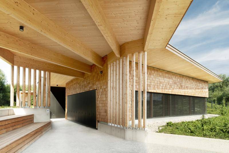 Das Gebäude mit seiner Holz-Schindelfassade und schwarzen Toren, sowie Jalousien vor den Fenstern. Der Vorplatz ist überdacht, ebenfalls mit einer Holzkonstruktion. Links sind Sitzflächen unter dem Dach angeordnet. Das Gebäude mit seiner Holz-Schindelfassade und schwarzen Toren, sowie Jalousien vor den Fenstern. Der Vorplatz ist überdacht, ebenfalls mit einer Holzkonstruktion. Links sind Sitzflächen unter dem Dach angeordnet.