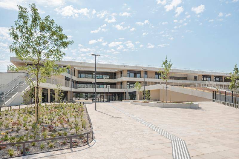 Zu sehen ist der Eingangsbereich des Gebäudes. Links im Bild leicht verdeckt durch einen jungen Baum. Das Gebäude besteht aus drei Stockwerken. Die Fassade ist in hellem Beige ausgefüht und die großen Fensterflächen sind mit grauen Rahmen eingefasst. Rechts steigt vor dem Gebäude das Gelände bis ins erste Stockwerk an. Zu sehen ist der Eingangsbereich des Gebäudes. Links im Bild leicht verdeckt durch einen jungen Baum. Das Gebäude besteht aus drei Stockwerken. Die Fassade ist in hellem Beige ausgefüht und die großen Fensterflächen sind mit grauen Rahmen eingefasst. Rechts steigt vor dem Gebäude das Gelände bis ins erste Stockwerk an.